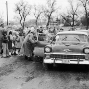 Children stand on a sidewalk beside cars after an evacuation at Lakewood Elementary School in Lakewood (Jefferson County), Colorado. Children and teachers or parents wear rain coats and scarves or hats. A license plate reads: "D11 3818, Colorado, '57."