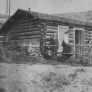 Log cabin residence of Martha and Mary Crane, Cripple Creek, Colorado; one woman sits on upper part of split rail fence, another woman stands near fence gate and one man stands in front of cabin; former residence of Ralph Carr, ex-Governor of Colorado is in the background; residence includes stone fence or foundation, low gable roof with shingles,  single entry at center, opened screen and front door, two double-sash plain windows; background includes outbuildings, mine tailings and surface buildings.