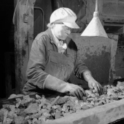 A woman worker sorts through rocks on a conveyor belt at a mill building for a London Mine near Alma (Parker County), Colorado. She wears work overalls, heavy gloves, and a cap. Glasses rest on her nose.