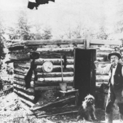 "Old Man Topping" with a miner's pick over his shoulder and his dog pose in front of first log cabin in Cripple Creek, Colorado; the cabin is small, flat-roof and rounded with a gold pan on exterior cabin wall and a shovel propped against cabin wall.