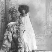 Studio portrait of a woman in a nightshirt or undergarment; she has a frizzy hair style and wears a cameo choker.