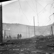View of Bennett Avenue's desolation and total destruction caused by second fire of 1896, Cripple Creek, Colorado; destruction includes burnt telegraph or utility poles and scattered debris of timbers; groups of men survey remains and destruction; Mount Pisgah is on distant horizon.