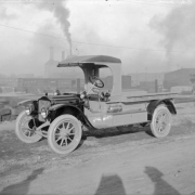 View of a truck used by the Mountain State Telephone and Telegraph Company in Pueblo (Pueblo County), Colorado. The truck has a hand brake, sirens, and plastic windshield rolled and attached to the roof of the automobile. Words painted on the side of the truck read: "Mountain States Tel. & Tel. Co." Smoke emerges from the smokestack of the Colorado Fuel and Iron Company steel mill in the distance.