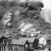 View from junction of Myers and "A" Streets across rooftops during April 29th, 1896 fire, Cripple Creek, Colorado; huge black clouds of smoke billow in the sky; scene includes one and two-story wooden frame and brick, gable and flat-roof residences and commercial buildings; horse-drawn wagons loaded with personal belongings are parked in front of small wooden frame residences (actual cribs of infamous red-light district of Myers Avenue).