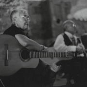 Unidentified musicians, members of the Guardiantes de Cultura pose in the Concilio Superior of La Sociedad Protection Mutua de Trabajadores Unidos meeting hall in Antonito (Conejos County), Colorado. An unidentified woman sits and holds a guitar, she wears eyeglasses and has gray hair, a man is beside her.