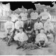 Photograph of a Elyria's baseball team.  The team sits in three rows, two on the ground, five on a bench, and four standing behind.  All wear their uniforms.  The upper portion of the photograph has damage and some of the faces can not been seen