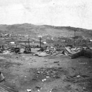 View of destruction caused by 1896 fire, Cripple Creek, Colorado; scene includes remains of twisted sheet metal, ashes and burnt timbers, and former business and commercial district; canvas tents are scattered amidst fire debris; remaining buildings and residences are in the distance on the hillside.