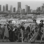 Members of a Chicano theater group identified as the National Chicano Theater, directed by Enrique Montano, pose on a rise looking over the skyline of Denver, Colorado.  They lean against or stand near two automobiles from the 1940's.  The men are dressed as Zoot suiters and the women wear vintage style dresses and skirts and blouses. Two men wear long jackets, pegged pants,  long chains and hats.