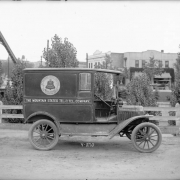 View of a Ford delivery truck used by the Mountain States Telephone and Telegraph Company in Denver, Colorado. A bell logo is painted on the side of the truck and lettering reads: "Mountain States Tel. & Tel. Company, Local and Long Distance Telephone, Bell System." Businesses and bridges are in the distance.
