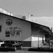 View of the Two Mule Inn at 500 Denargo Market in the Five Points neighborhood of Denver, Colorado.  This former produce warehouse is made of brick and has a metal gabled roof.  Signs read: "Two Mule Inn," "Budweiser," and "Welcome Colorado Rockies Fans." Skyscrapers are in the distance.