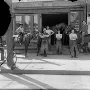 A group of men pose near possibly a blacksmith's shop in Denver, Colorado. Two men are on horseback and other men wear long aprons.