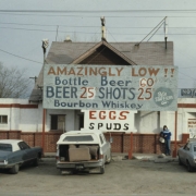 A woman poses in front of Dan's Corner Tavern,  5302 Brighton Boulevard in the Elyria-Swansea neighborhood of Denver, Colorado. The bar, a former residence, has a cross-gabled roof, brick facing and several additions. Signs read, "AMAZINGLY LOW!!, Bottle Beer 60 cents, BEER 20 cents, SHOTS 25 cents, Bourbon Whiskey, Pete Staffieri," "EGGS, SPUDS," "NOTARY PETE" and "Notary Public, Bottle Beer." A phone booth is near the building and cars are parked nearby.