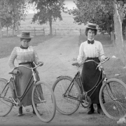 Women pose with their bicycles in a park possibly in Denver, Colorado. They wear dresses and straw wide-brimmed hats.