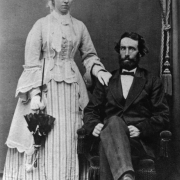 Studio wedding portrait of Mr. Isaac Watson Wallace and his bride Henrietta McHeffey Wallace; she wears a striped skirt, bodice, long jacket, flared cuffs, gloves, tulle headpiece, and carries a parasol.