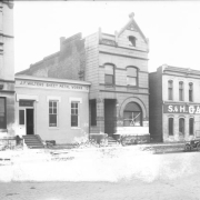 Exterior view of the Tri-State Buddhist Temple (Church) at 1942 Market Street in downtown Denver, Colorado. The multi-story, brick building has a rounded arch window on the first floor facade, a gabled parapet with finial and roof battlements. Signs on additional buildings read: "S.& H. Garage Repair" and "J.F. Walters Sheet Metal Works." An automobile is parked on the street.