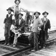 Five Japanese workers pose around a railroad handcart. Each wears  workclothes, including heavy boots, hats, and gloves.