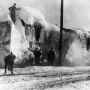 Four Denver & Salt Lake Railroad workers stand next to and on a snow-encrusted rotary plow that was involved in a train accident in Tabernash, Colorado in Grand County.  Steam rises off of the engine, and snow covers the ground around the tracks.