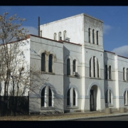 View of the State Armory (1889; William Quayle) at 2565 Curtis Street in the Five Points neighborhood of Denver, Colorado. Shows a two-story, brick, Romanesque Revival influenced, symmetrical building with semi-circular arched windows, belt courses and an inset entrance in a three-story tower. The brick walls have been stuccoed. The former armory is part of the Curtis Park-Champa Street Historic District and now houses Scientific Site Inc.
