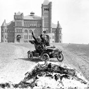 Harry M. Rhoads, behind the steering wheel, poses in a convertible with passenger in front of Westminster University or College, at 3455 West 83rd Avenue in Westminster (Adams County), Colorado. Animals graze in the barren field in which the building is situated. Architectural features of the building: sandstone construction, 175-foot tower with a hipped roof, conical towers, square towers, arched and square windows and entrances, gables, and chimneys.