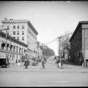 View east down 16th (Sixteenth) Avenue from Broadway, Denver, Colorado; several early automobiles on street & parked at curb, horse-drawn wagon, man riding bicycle; Denver Auto Goods Company (1600 or 1614 Broadway), Young Men's Christian Association building, Johnson's Prescriptions (or Johnson Drug Company, Joseph S. Johnson, druggist, 1572 Broadway); men on bicycles, enclosed carriage or coach, barber pole, mail box, fire hydrant.