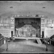 Interior view of El Jebel Temple, Denver, Colorado; local Masonic order of El Jebel Shriners (building dedicated November, 1907); shows stage area of auditorium with chairs and bench on stage, backdrop, curtain draped open, wall mural painted by Jens Eriksen, ceiling with light bulbs arranged to depict stars, upright piano, radiators, and potted palm trees. The temple was designed by the Baerresen Brothers.