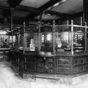 Interior view of the First National Bank, Colorado Springs, El Paso County, Colorado, features coffered wood ceilings and an ornate, three-windowed tellers box and brass footrails under writing shelves. A. H.  Hunt, the cashier is behind beaded grating and another man stands in the background. Signs read: "Cashier," "Teller," "Next Window," and "Revenue Stamps for sale at This window."