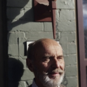 William Allen West, author and historic preservation advocate, poses on the porch of his home in the Curtis Park-Champa Street Historic District, in the Five Points neighborhood, Denver, Colorado. He is balding, has a beard and mustache and wears a sweater.