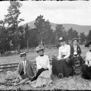 Group portrait of a man, women, and a boy at a picnic in Colorado; costume includes hats with flowers. Hills and railroad tracks are in the background.