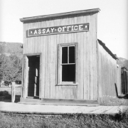 View of the dilapidated Assay Office in Pitkin, Gunnison County, Colorado, a wooden, false front building with vertical board siding and a plank sidewalk surrounded by unkempt weeds and grass.