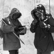 Two AdAmAn club members stand in front of a ice wall with icycles and prepare for the annual New Year's Eve Pikes Peak climb, Colorado Springs, El Paso County, Colorado. The men wear parkas and mittens; one has a backpack with an antenna, the other holds an ice- climbing pick, a "Motorola," portable radio and a coiled rope.