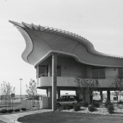 View of the Centennial Airport  terminal building at 7800 South Peoria Street in Englewood (Arapahoe County), Colorado. The wing shaped entrance of the airport has a drive way wrapping around the exterior. A  runway is in the background. A tall column supports the underside of the wing, which curves up to the edges that are lined with metal protrusions which represent feathers. The building was constructed in 1967.
