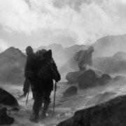 AdAmAn Club members climb among boulders, ice, and blowing snow, on Pikes Peak, Colorado Springs, El Paso County, Colorado. They wear parkas, backpacks, and have walking sticks.
