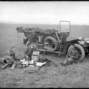 Three men picnic next to an early gasoline-powered automobile near dam site for Standley Lake, Colorado; men eat apples and sit on car seat cushions; scene includes blanket, wicker picnic basket, food items, plates, bottles, a spare tire attached to automobile runner, and the phographer's box of supplies in backseat. McClure photographed construction of Standley Lake by Kenefick-Quigley-Russel Construction Company.