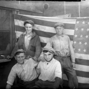 Group portrait of teenaged boys by a United States flag in Denver, Colorado; costume includes duster hats, a knit sweater, and a cigarette.