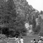 Grace Noel, a young girl, casts a line into South Boulder Creek in Eldorado Canyon State Park in Boulder County, Colorado.  People are near her on the edge of the creek. A child is on the opposite bank. The jagged rock outcrops of the canyon walls are in the distance.