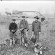 Teenaged boys with axes pose by Archer Canal in Denver, Colorado. A dog and a man with a bicycle are nearby. A horse-drawn wagon makes its way over the Alameda Avenue bridge in the distance.