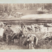 Miners pose with a pack train of burros near a lake or pond in the Red Mountain Mining District in Ouray or San Juan County, Colorado.  Two of the men are African American. The men wear hats, and two smoke pipes. The burros carry split wooden logs. Shows a wooden post  in a cairn.