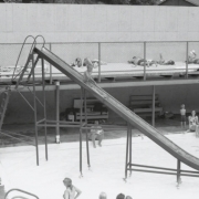 View of the pool at Eldorado Springs Resort in Eldorado Springs (Boulder County), Colorado. A child goes down a tall water slide.  Children are lined up to use the slide.  The pool has a sun deck over a shade area. The resort was originally named Moffat Lakes Resort,  and when it opened (1906) it was advertised as the largest swimming pool in the country.