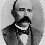 Portrait of semi-bald man with a "walrus" moustache, wearing a jacket and overcoat with wide lapels. His bow tie is adorned with a star- shaped pin.