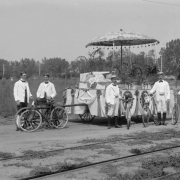 Men in white jackets, kneepants, and dusters pose with their Sunflower Carnival Parade float and bicycles, Colorado Springs, El Paso County, Colorado. The float is decorated with a patterned, tasseled, paper parasol, bunting, flowers, potted palms and date: "1897." Streetcar tracks are in the foreground.