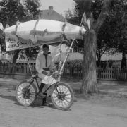 An entrant in the Sunflower Carnival parade poses on a bicycle in  his white shirt and kneepants, Colorado Springs, El Paso County, Colorado.   His bicycle is decorated with white fabric woven into the spokes, a dirigible blimp with propellers mounted on poles, fringe, a United States flag and sign: "I Will Jump Sunday Nit."
