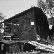 A dilapidated log cabin in Liberty, Saguache County, Colorado, has a bench on the porch, and a ramp. Bird wings are next to the door, by a  sign: "Notice."