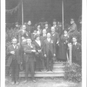 A group of men and women stand on the steps and the porch of a house in probably Mexico City, Mexico. Standing bottom left is José Jesús Córdova, County Commissioner for Las Animas County Colorado, he wears a suit, vest and tie and has his hand in his vest. he has a full mustache. Center on the first step is Colorado State Senator and Mexican Consul Casimiro Barela. The elderly Senator Barela wears a suit vest and tie and holds a hat. He has gray hair and a mustache.