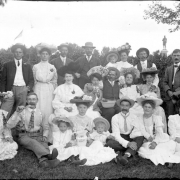 Outdoor portrait of a group of men, women, and children in City Park, Denver, Colorado. One woman holds a pitcher while another eats from a bowl. A statue of Robert Burns is in the distance.