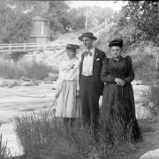 Outdoor portrait of a man and two women near a creek in the mountains of Colorado. The women wear hats decorated with flowers. A water tank and a wooden bridge are in the distance.