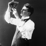 Self portrait of George Lytle Beam examining a glass plate. He wears a lab apron, striped bow tie, white shirt with wing-tip collar and his pince-nez eyeglasses.