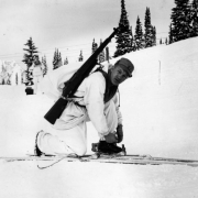 Tenth Mountain Division Corporal, Richard Cochran bends over his skis as though adjusting his bindings. He is wearing winter camouflage of "whites" as well as a knapsack and his rifle. Almost obscured behind him is  another soldier in the same uniform standing and facing directly toward the camera. Both are on a snow covered mountain in an unnamed location.