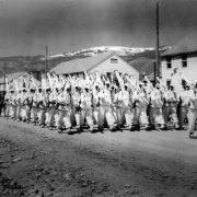 10th Mountain Division soldiers parade down a street at Camp Hale, Colorado. They are wearing their "whites" the winter camouflage uniforms and carry white skis on their right shoulder as rifles are normally carried while on parade.