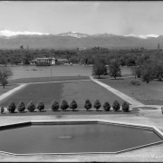 View of range of Rocky Mountains, including 14,260-foot Mount Evans at left background, from City Park in Denver, Colorado; shows octagonal fountain pool, landscaped lawn with pedestrians on pathway, lake with pavilion and bandshell, and the skyline of downtown Denver is visible.