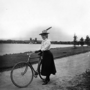 Woman with a bicycle at City Park, Denver, Colorado, wears gloves, a locket, low-heeled shoes, and a hat with a flower and feather. A pavilion and lake are in the background.
